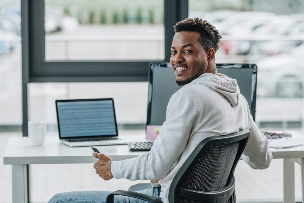 happy african american programmer smiling at camera while sitting at workplace in office